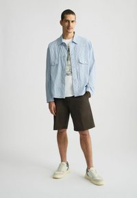 Cămașă cu dungi albastre deschise și nasturi, tricou alb cu grafică, pantaloni scurți maro și adidași neutri. Modelul stă în fața unui fundal simplu.