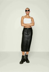 Saia midi de couro preto com costura frontal e detalhes de botão prateado, combinada com um top cropped branco e botas pretas com atacadores.