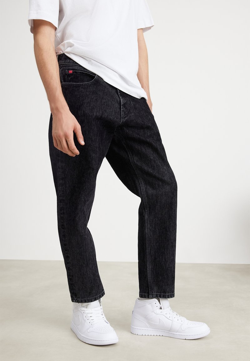 Homme portant un jean noir, des baskets blanches et un t-shirt blanc, debout devant un fond clair uni.