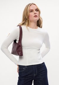 Biała bluzka z długim rękawem o dopasowanym kroju i szwie w talii, zestawiona z burgundową skórzaną torebką z plecionymi akcentami i miękką fakturą.
