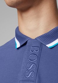 Polo shirt blu navy con colletto a costine caratterizzato da strisce turchesi e bianche. Logo ricamato con la scritta "BOSS" sulla patta. Texture morbida.