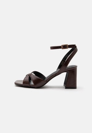 Sandale marron en cuir à talon haut, avec bride à la cheville, motif croisé à l’avant et talon bloc sur un fond clair uni.