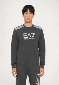 Tmavošedá mikina s dlhými rukávmi, rebrovaným výstrihom a bielym logom "EA7 Emporio Armani". Obsahuje pruhované akcenty na ramenách.