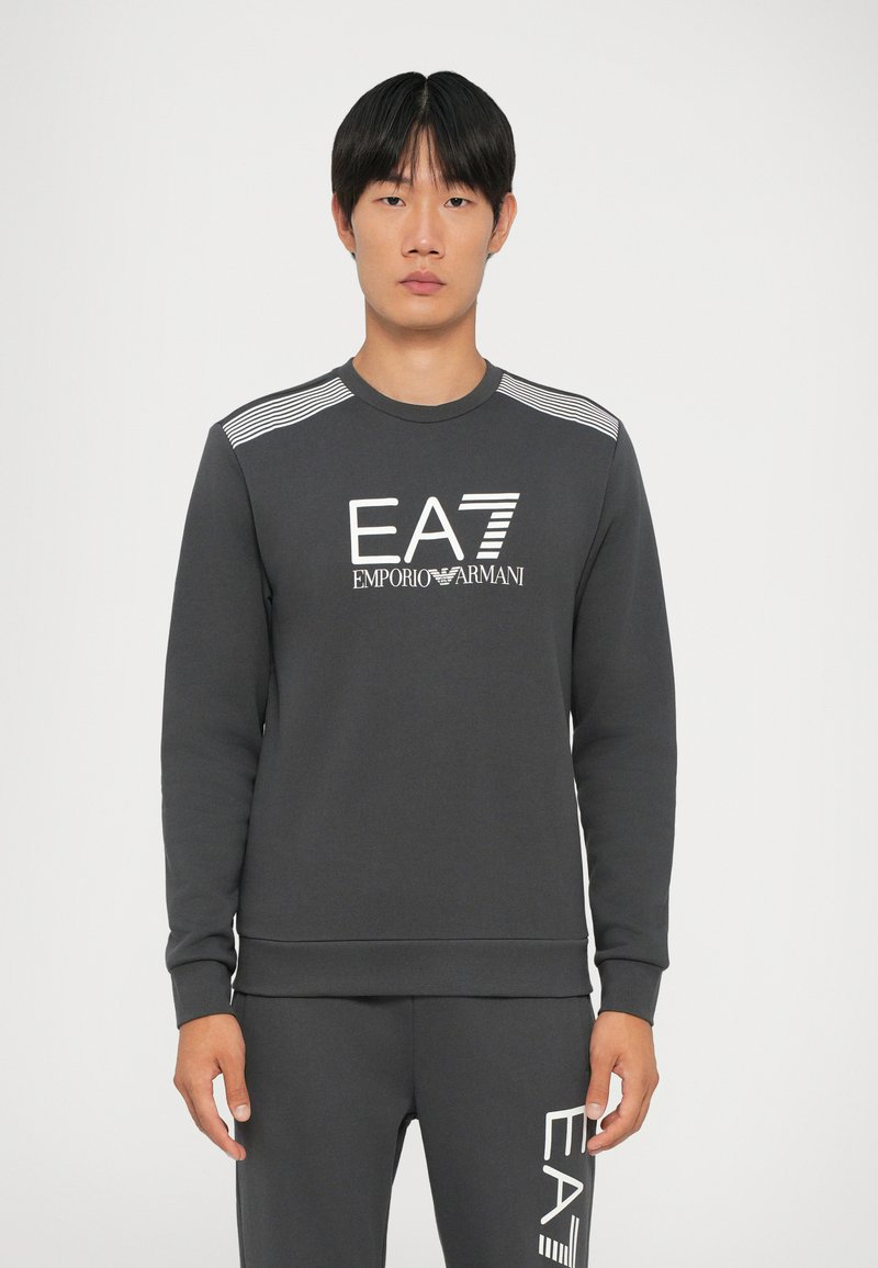 Tmavošedá mikina s dlhými rukávmi, rebrovaným výstrihom a bielym logom "EA7 Emporio Armani". Obsahuje pruhované akcenty na ramenách.