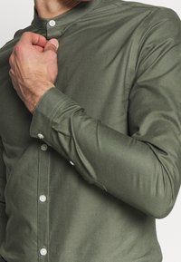 Oliivinvihreä napillinen paita ilman kauluksetta, pitkähihaiset hihat ja sileä pinta. Rintapellissä ja hihansuissa valkoiset napit.