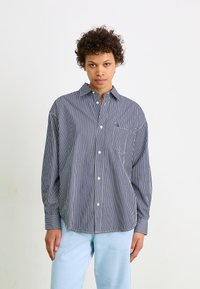 Chemise à manches longues à rayures bleues marines et blanches, avec une coupe décontractée et une poche poitrine. Pantalon bleu clair complétant le look.