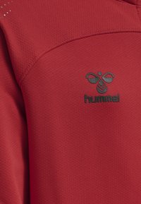 Rotes Sportshirt aus strukturiertem Stoff mit schwarzem Logo und Branding auf der Brust. Das Design umfasst subtile Mesh-Details.