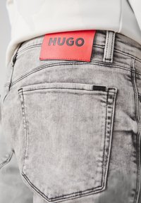 Szare spodnie dżinsowe mają czerwoną skórzaną etykietę na pasku z wytłoczonym czarnym napisem "HUGO". Tkanina ma wyblakłą fakturę i klasyczny design kieszeni.