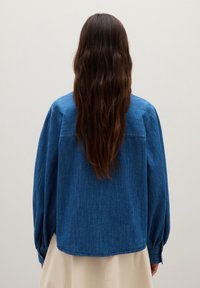 Person with long dark hair wearing a loose blue denim jacket and a beige skirt, facing away against a plain light background.