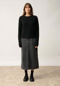 Pull en tricot noir avec deux poches avant, associé à une longue jupe en denim grise. Des chaussures noires épaisses complètent le look. Fond uni.