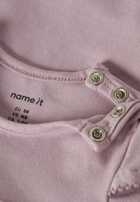 Helles rosa Kurzarmhemd mit silbernen Druckknöpfen, aufgedruckten Größenetiketten und einer weichen, glatten Baumwollstruktur. Die Kanten sind genäht.