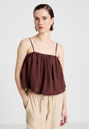 Femme portant un haut marron ample sans manches avec des bretelles fines et un pantalon beige clair à cordon contre un fond uni.