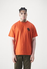 Camiseta de algodón naranja con mangas cortas y cuello redondo, que presenta un pequeño diseño gráfico negro en la zona izquierda del pecho.