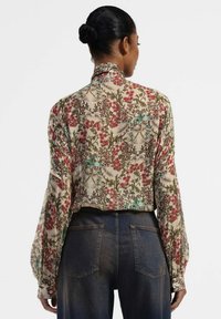 Blusa floreale con sfondo crema e motivi rossi, verdi e blu. Maniche lunghe con polsini elastici. Abbinata a jeans blu sbiaditi.