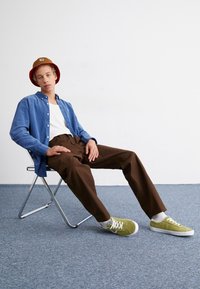 Blue button-up shirt, white tank top, brown pants, green sneakers, and a red bucket hat. Chair is metallic. Soft-textured floor.