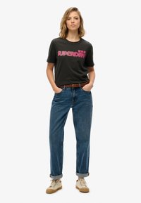 Czarna bawełniana koszulka z różowym napisem "SUPERDRY", zestawiona z niebieskimi dżinsami o wysokim stanie i beżowymi sneakersami z podwiniętymi rękawami.