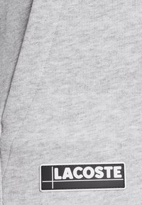 Tissu gris avec une étiquette rectangulaire noire et blanche affichant le mot LACOSTE en lettres majuscules grasses.