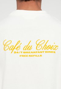 White cotton t-shirt featuring yellow text on the back. Text includes "Café du Choix," "24/7 Breakfast Diner," and "Free Refills."