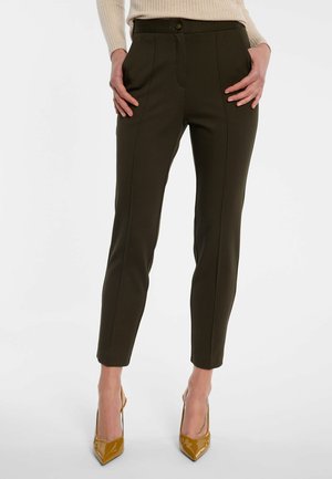 Vrouw draagt donker olijfgroene slim fit broek en mosterdgele hoge hakken, staand met handen bij de zakken tegen een effen achtergrond.