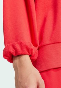 Červená mikina z látky s textúrovaným dizajnom manžiet; vrúbkovaná textúra na zápästí; má voľný strih a spodný okraj, ktorý zdôrazňuje hebkosť materiálu.