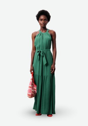 Femme portant une robe maxi verte sans manches avec un col halter et une taille nouée, tenant un sac rose à motifs et portant des chaussures rouges.