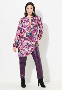 Blusa colorata con motivo astratto in rosa, viola e blu. Presenta un colletto a V e maniche lunghe. Abbinata a pantaloni aderenti in finta pelle viola.