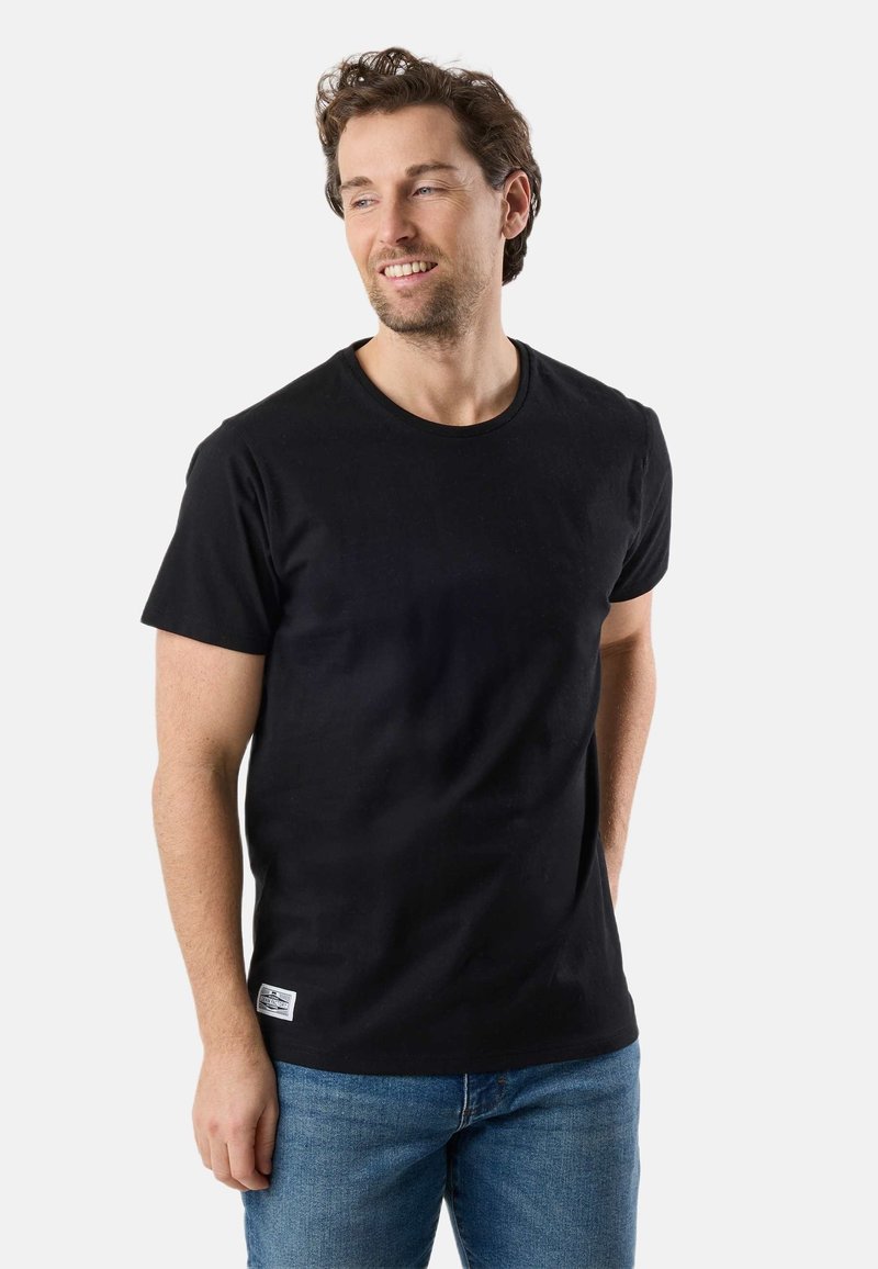 Homme aux cheveux bruns courts et bouclés, souriant et regardant vers la droite, portant un t-shirt noir et un jean bleu, debout devant un fond uni.
