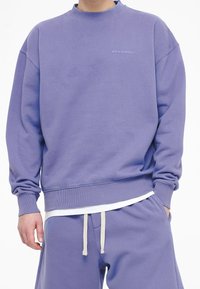 Uomo che indossa una felpa viola e pantaloni della tuta abbinati con coulisse bianche, in piedi con una mano in tasca.