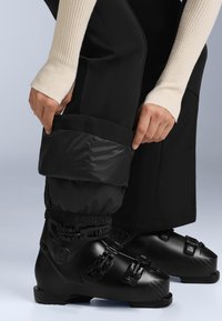 Fekete sícipők négy állítható csattal, fekete párnázott sínadrággal, elasztikus mandzsettával, világos háttér előtt.