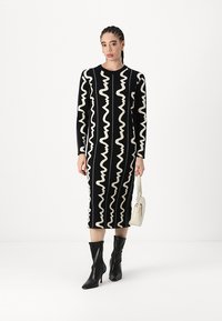 Robe en tricot avec un motif ondulé noir et blanc, des manches longues et une longueur midi. Associée à des bottes à talons noires et un sac à main blanc.