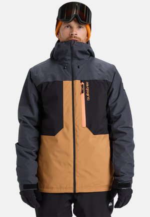 Homme portant un bonnet orange, des lunettes de ski noires, une veste de snowboard noire et marron, des gants noirs et un pantalon noir devant un fond clair.