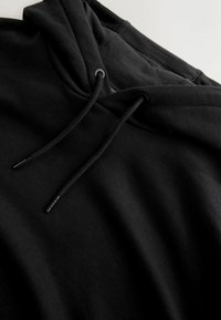 Czarna bluza z kapturem o miękkiej fakturze, z dekoltem ze sznurkiem i oczkami. Tkanina wydaje się gruba i gładka, z luźnym krojem.