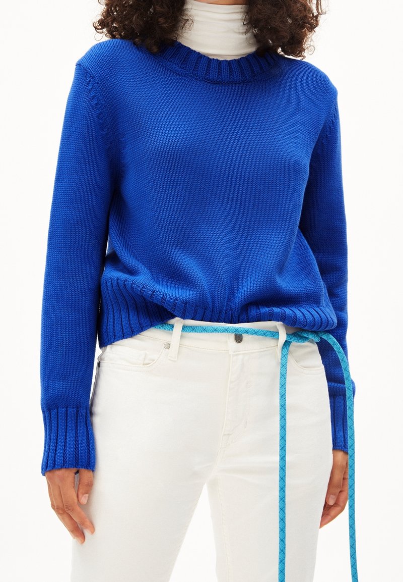 Personne portant un pull en maille bleu vif sur un col roulé blanc, un pantalon blanc, et une longue ceinture bleue en corde pendue, non nouée à la taille.