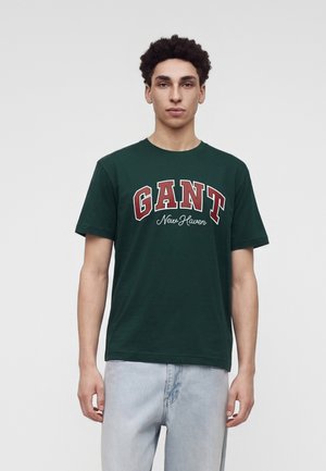 Jeune homme aux cheveux bouclés portant un t-shirt GANT vert foncé et un jean bleu clair, debout devant un fond clair uni.