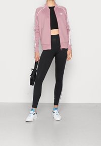 Růžová mikina na zip s bílými pruhy, černý crop top, černé legíny s bílými bočními pruhy a bílé tenisky s modrými akcenty.