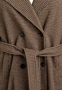 Hnedý kabát s houndstooth vzorom, dvojitým zapínaním, veľkými čiernymi gombíkmi a zodpovedajúcim látkovým opaskom. Textúrovaná tkanina.