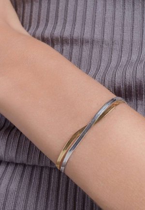 Bracciale a strati in oro e argento con filamenti testurizzati, caratterizzato da un design flessibile che si adatta al polso su uno sfondo grigio a righe.