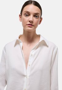 Chemise blanche à boutons en tissu léger avec un col pointu et des manches longues, présentant un décolleté en V profond et des boutons en forme de perle.