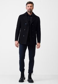 Zwarte wollen peacoat met een dubbel-breasted ontwerp, grote kraag, blauwe knopen, gedragen over een zwart shirt en marineblauwe broek, gecombineerd met zwarte laarzen.