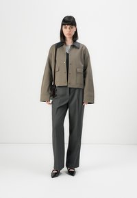 Jonge vrouw met een zwart bobkapsel, gekleed in een olijfgroene jas met een zwarte kraag, grijze broek, zwarte puntige schoenen en een donkere schoudertas.