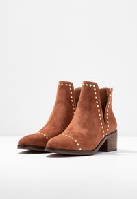 Bottines en daim marron avec un haut courbé, des clous dorés, et un petit talon en bois, présentant une texture lisse et un design à bout pointu.