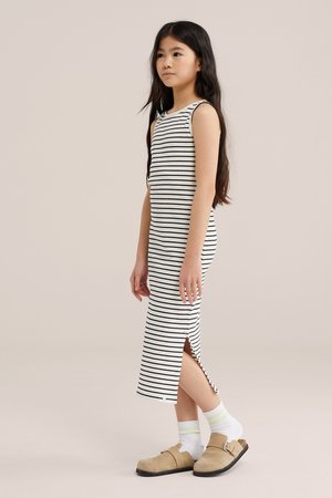 Mladé dievča stojace bokom, ktoré má na sebe biele šaty bez rukávov s čiernymi pruhmi, biele ponožky a béžové slip-on topánky.