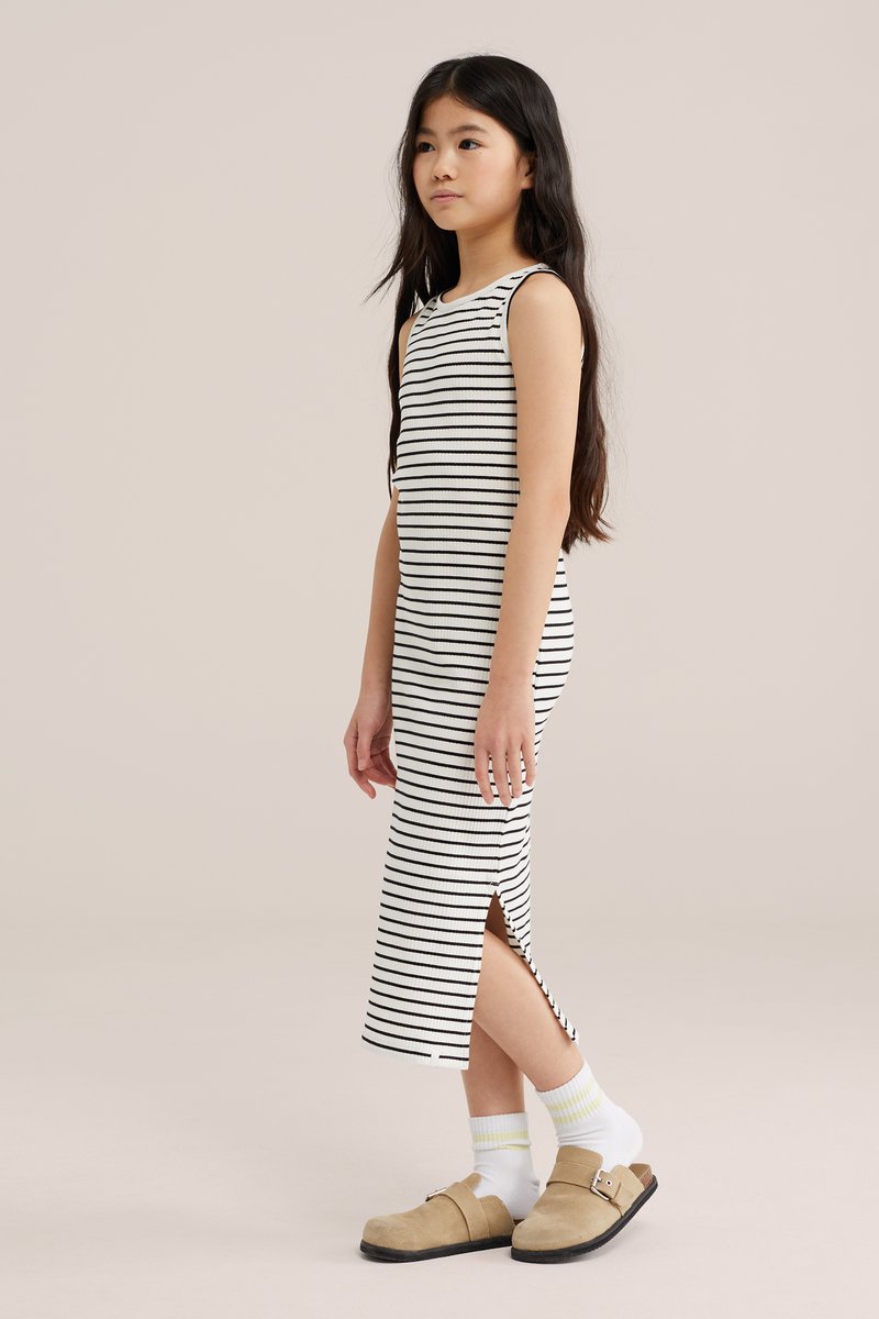 Jeune fille debout de profil portant une robe blanche sans manches avec des rayures noires, des chaussettes blanches et des chaussures beiges sans lacets.