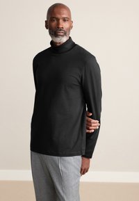 Sort langærmet rullekraveskjorte lavet af glat stof, med høj krave og løs pasform, kombineret med lysegrå bukser.