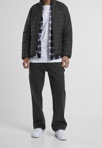 Black quilted jacket over a white t-shirt and checked flannel shirt. Worn with dark cargo pants and white sneakers. Visible zipper and pockets.