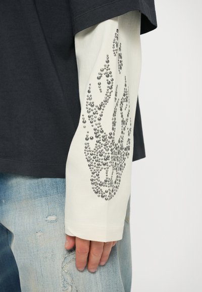 Camicetta nera a maniche corte indossata sopra una camicia bianca a maniche lunghe, con decorazioni di perline argentate e jeans in denim consumati.