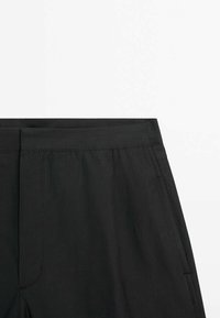 Czarne spodnie z płaskim pasem, z przednimi kieszeniami i gładką teksturą. Materiał wydaje się lekki i trwały.