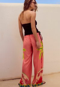 Pantalons larges roses avec un motif de palmiers jaunes et verts. Présentent une coupe décontractée et une taille élastique avec cordon de serrage.