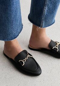 Černé kožené nazouvací boty se špičatou špičkou a zdobené zlatým řetízkem. Nošené s ohrnutými modrými džínami. Plochá podrážka a hladká textura.