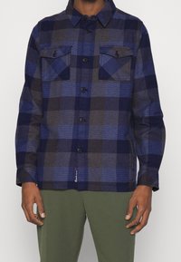 Koszula flanelowa w kratę w odcieniach granatu i niebieskiego, z dwiema kieszeniami na piersi, zapięciem na guziki i luźnym krojem. Wykonana z fakturowanej tkaniny.